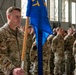 A U.S. Air Force military formation ceremony taking place inside a hangar. A senior non-commissioned officer identified by name tape as 'RAU' stands at the forefront holding a blue unit guidon flag with gold insignia. Multiple Airmen in OCP (Operational Camouflage Pattern) uniforms are standing at attention in formation behind him.
