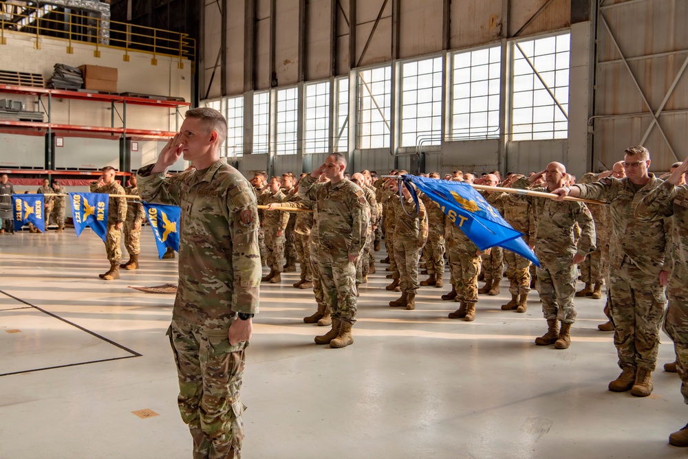 A large formation of military personnel in OCP (Operational Camouflage Pattern) uniforms are rendering a salute inside a large aircraft hangar. The ceremony appears to be a formal military formation event, likely a change of command, activation, or similar official ceremony. Blue unit guidons with gold insignia are visible throughout the formation.