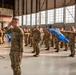 A large formation of military personnel in OCP (Operational Camouflage Pattern) uniforms are rendering a salute inside a large aircraft hangar. The ceremony appears to be a formal military formation event, likely a change of command, activation, or similar official ceremony. Blue unit guidons with gold insignia are visible throughout the formation.