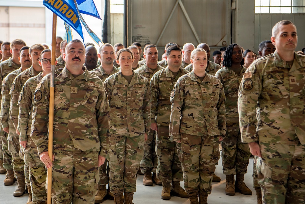 A large formation of U.S. Air Force personnel in OCP (Operational Camouflage Pattern) uniforms standing at attention inside what appears to be a hangar facility. One service member in the front row is holding a blue squadron guidon flag. The formation appears to be assembled for an official military ceremony or unit formation.