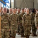 A large formation of U.S. Air Force personnel in OCP (Operational Camouflage Pattern) uniforms standing at attention inside what appears to be a hangar facility. One service member in the front row is holding a blue squadron guidon flag. The formation appears to be assembled for an official military ceremony or unit formation.