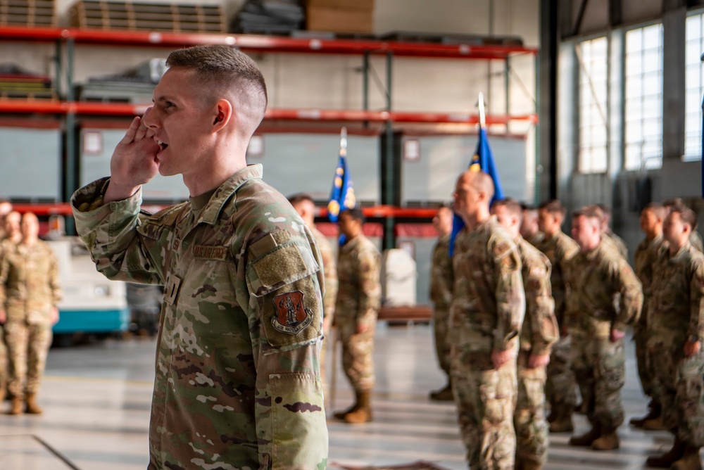 A U.S. Air Force service member in the foreground is shouting commands with his hand cupped near his mouth during what appears to be a formal military formation ceremony inside a large hangar or warehouse facility. Multiple personnel in OCP (Operational Camouflage Pattern) uniforms are standing in formation in the background. Blue Air Force guidon flags are visible among the assembled troops.