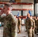 A U.S. Air Force service member in the foreground is shouting commands with his hand cupped near his mouth during what appears to be a formal military formation ceremony inside a large hangar or warehouse facility. Multiple personnel in OCP (Operational Camouflage Pattern) uniforms are standing in formation in the background. Blue Air Force guidon flags are visible among the assembled troops.
