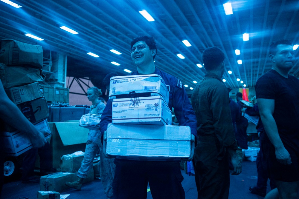 A smiling service member in a blue coverall uniform carries a stack of USPS Priority Mail packages inside what appears to be a large military vessel's hangar bay or cargo hold. The space is illuminated by blue-tinted fluorescent lighting and is filled with other personnel and packages. The scene appears to depict a mail call or logistics operation aboard a naval ship.