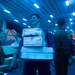 A smiling service member in a blue coverall uniform carries a stack of USPS Priority Mail packages inside what appears to be a large military vessel's hangar bay or cargo hold. The space is illuminated by blue-tinted fluorescent lighting and is filled with other personnel and packages. The scene appears to depict a mail call or logistics operation aboard a naval ship.