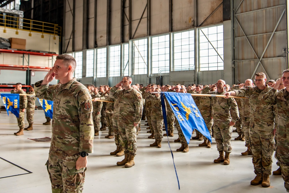 A large formation of military personnel in OCP (Operational Camouflage Pattern) uniforms are rendering a salute during a formal ceremony inside a military hangar. Multiple unit guidons (blue flags with gold insignia) are being held horizontally by designated guidon bearers. The ceremony appears to be a formal military event such as a change of command, activation, or deployment ceremony.