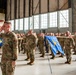 A large formation of military personnel in OCP (Operational Camouflage Pattern) uniforms are rendering a salute during a formal ceremony inside a military hangar. Multiple unit guidons (blue flags with gold insignia) are being held horizontally by designated guidon bearers. The ceremony appears to be a formal military event such as a change of command, activation, or deployment ceremony.
