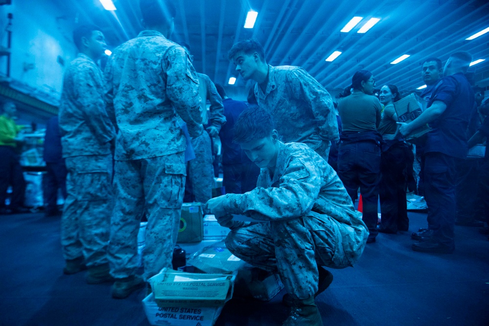 Military personnel in camouflage uniforms are gathered in what appears to be a ship's hangar bay or cargo hold, illuminated by a strong blue light. Several service members are sorting through United States Postal Service packages and boxes on the floor. The scene suggests a mail call or supply distribution event aboard a naval vessel.