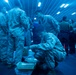 Military personnel in camouflage uniforms are gathered in what appears to be a ship's hangar bay or cargo hold, illuminated by a strong blue light. Several service members are sorting through United States Postal Service packages and boxes on the floor. The scene suggests a mail call or supply distribution event aboard a naval vessel.