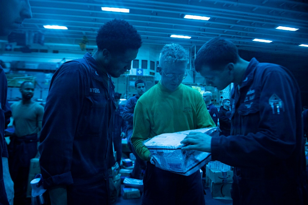 Military personnel, likely U.S. Navy sailors, are gathered in what appears to be a ship's interior bay or hangar deck under blue lighting. Three individuals in the foreground are examining a package or parcel, with one wearing a green shirt and another with a name tag reading 'MYERS'. Additional personnel and stacked packages are visible in the background, suggesting a mail or supply distribution operation.