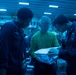 Military personnel, likely U.S. Navy sailors, are gathered in what appears to be a ship's interior bay or hangar deck under blue lighting. Three individuals in the foreground are examining a package or parcel, with one wearing a green shirt and another with a name tag reading 'MYERS'. Additional personnel and stacked packages are visible in the background, suggesting a mail or supply distribution operation.