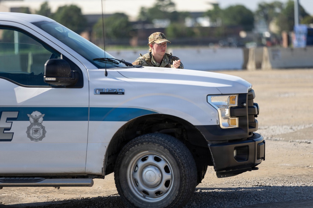 A military or security personnel member in camouflage uniform and cap is positioned behind a white Ford F-150 XL pickup truck marked with what appears to be a US Air Force Security Forces emblem and blue striping. The individual appears to be in a tactical or observational stance, looking to the side. The setting appears to be a gravel or dirt area, possibly a military base or secured perimeter.