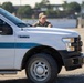 A military or security personnel member in camouflage uniform and cap is positioned behind a white Ford F-150 XL pickup truck marked with what appears to be a US Air Force Security Forces emblem and blue striping. The individual appears to be in a tactical or observational stance, looking to the side. The setting appears to be a gravel or dirt area, possibly a military base or secured perimeter.