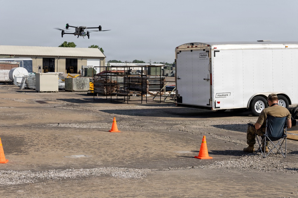 A person in military-style clothing sits in a folding chair operating a drone in an industrial or military yard setting. The quadcopter drone is airborne in the upper left portion of the frame, while orange traffic cones mark a designated flight area on the gravel surface. A white Haulmark enclosed trailer and various storage structures are visible in the background.