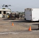 A person in military-style clothing sits in a folding chair operating a drone in an industrial or military yard setting. The quadcopter drone is airborne in the upper left portion of the frame, while orange traffic cones mark a designated flight area on the gravel surface. A white Haulmark enclosed trailer and various storage structures are visible in the background.