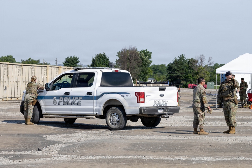 The image shows military personnel in camouflage uniforms interacting around a white police pickup truck (Ford F-150) marked with 'POLICE' and 'DIAL 9-1-1' in an outdoor staging area. Several soldiers are positioned near the vehicle and in the background near a white canopy tent. The setting appears to be a military or joint law enforcement exercise or operational staging area.