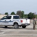 The image shows military personnel in camouflage uniforms interacting around a white police pickup truck (Ford F-150) marked with 'POLICE' and 'DIAL 9-1-1' in an outdoor staging area. Several soldiers are positioned near the vehicle and in the background near a white canopy tent. The setting appears to be a military or joint law enforcement exercise or operational staging area.