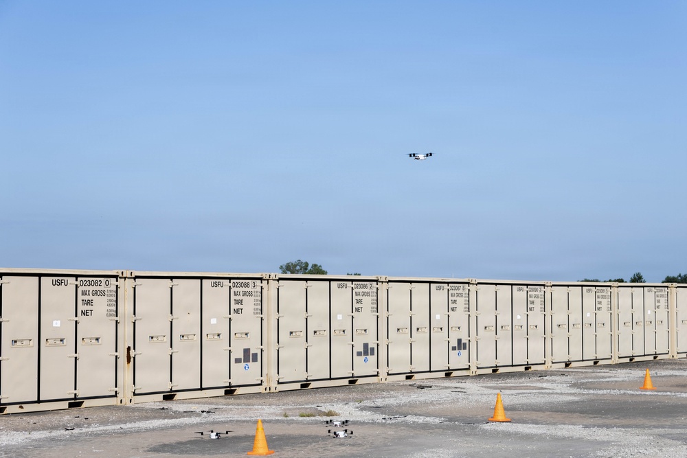 The image shows a row of tan/beige military-style shipping containers marked with 'USFU' prefixes and identification numbers, arranged in a long line on a gravel surface. Several small drones are visible on the ground near orange traffic cones, with at least one drone airborne above the containers. The scene appears to depict a drone testing or demonstration exercise at a logistics or storage facility.