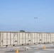 The image shows a row of tan/beige military-style shipping containers marked with 'USFU' prefixes and identification numbers, arranged in a long line on a gravel surface. Several small drones are visible on the ground near orange traffic cones, with at least one drone airborne above the containers. The scene appears to depict a drone testing or demonstration exercise at a logistics or storage facility.