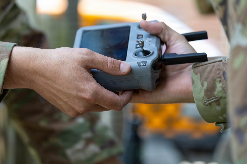Two individuals in military camouflage uniforms are handling a DJI drone remote controller with an integrated screen. The controller appears to be a DJI Smart Controller or similar model, featuring dual antennas, a built-in display, and control joysticks. The image captures a close-up handoff or collaborative inspection of the device.