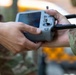 Two individuals in military camouflage uniforms are handling a DJI drone remote controller with an integrated screen. The controller appears to be a DJI Smart Controller or similar model, featuring dual antennas, a built-in display, and control joysticks. The image captures a close-up handoff or collaborative inspection of the device.