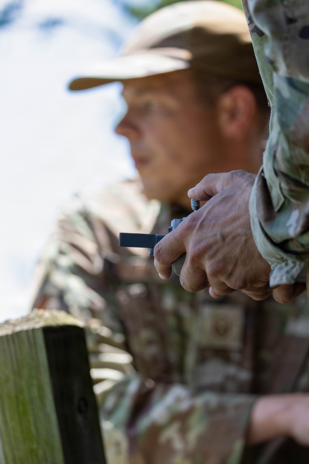 A person wearing camouflage clothing and a tan/camouflage cap is holding a small dark-colored device in their hands. The image is taken with a shallow depth of field, keeping the hands and device in sharp focus while the person's face is blurred in the background. The outdoor setting and camouflage attire suggest a hunting or military/tactical context.