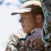 A person wearing camouflage clothing and a tan/camouflage cap is holding a small dark-colored device in their hands. The image is taken with a shallow depth of field, keeping the hands and device in sharp focus while the person's face is blurred in the background. The outdoor setting and camouflage attire suggest a hunting or military/tactical context.