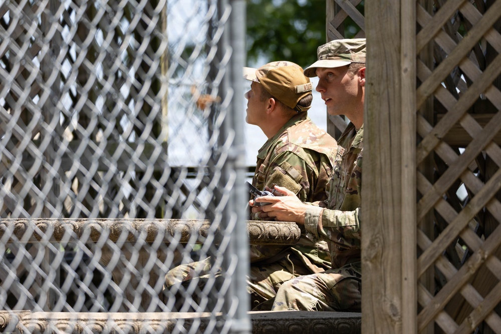 Two individuals in military camouflage uniforms (OCP/MultiCam pattern) are seated together in what appears to be an elevated wooden structure or observation platform. One person is holding a small device, possibly a remote control or radio, while both appear to be looking outward in the same direction. The scene is partially obscured by a chain-link fence in the foreground.