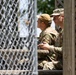 Two individuals in military camouflage uniforms (OCP/MultiCam pattern) are seated together in what appears to be an elevated wooden structure or observation platform. One person is holding a small device, possibly a remote control or radio, while both appear to be looking outward in the same direction. The scene is partially obscured by a chain-link fence in the foreground.