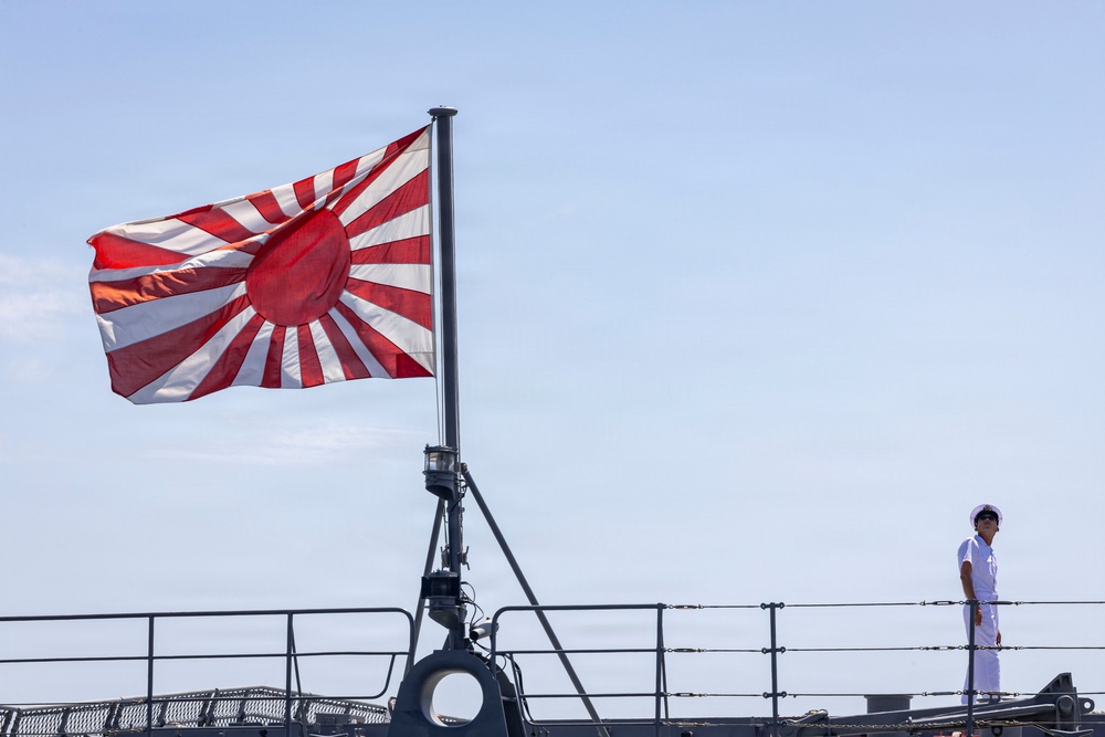 A naval vessel is shown with the Japan Maritime Self-Defense Force ensign (Rising Sun flag) flying prominently from a mast. A single crew member dressed in white naval uniform and white cap stands at attention on the deck. The image is taken from a low angle against a clear blue sky, emphasizing the flag and the sailor's silhouette.