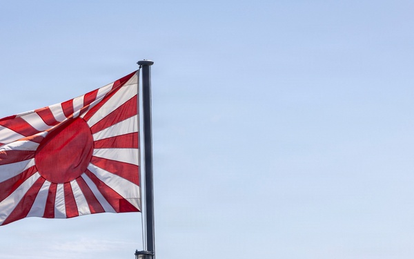 A naval vessel is shown with the Japan Maritime Self-Defense Force ensign (Rising Sun flag) flying prominently from a mast. A single crew member dressed in white naval uniform and white cap stands at attention on the deck. The image is taken from a low angle against a clear blue sky, emphasizing the flag and the sailor's silhouette.
