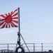 A naval vessel is shown with the Japan Maritime Self-Defense Force ensign (Rising Sun flag) flying prominently from a mast. A single crew member dressed in white naval uniform and white cap stands at attention on the deck. The image is taken from a low angle against a clear blue sky, emphasizing the flag and the sailor's silhouette.
