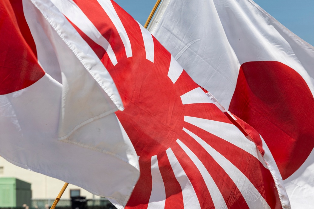 The image shows multiple Japanese flags flying together outdoors in what appears to be a sunny setting. Visible are both the standard Japanese national flag (Nisshōki/Hinomaru) featuring a red circle on white, and the Japanese Rising Sun flag (Kyokujitsu-ki) featuring a red sun with radiating rays on white. The flags are in motion, suggesting wind, and are photographed in close-up from below.