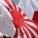 The image shows multiple Japanese flags flying together outdoors in what appears to be a sunny setting. Visible are both the standard Japanese national flag (Nisshōki/Hinomaru) featuring a red circle on white, and the Japanese Rising Sun flag (Kyokujitsu-ki) featuring a red sun with radiating rays on white. The flags are in motion, suggesting wind, and are photographed in close-up from below.