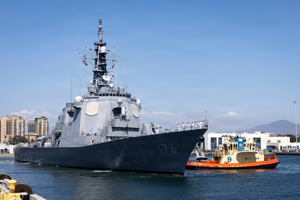 A large gray military warship bearing hull number 176 is maneuvering into port, assisted by an orange and yellow tugboat numbered 9. Crew members dressed in white uniforms are visible standing in formation along the ship's deck. The scene appears to be at a naval base with urban buildings and mountains visible in the background.