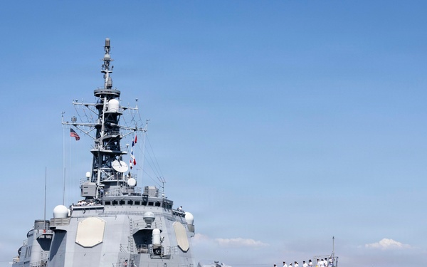 A large gray military warship bearing hull number 176 is maneuvering into port, assisted by an orange and yellow tugboat numbered 9. Crew members dressed in white uniforms are visible standing in formation along the ship's deck. The scene appears to be at a naval base with urban buildings and mountains visible in the background.