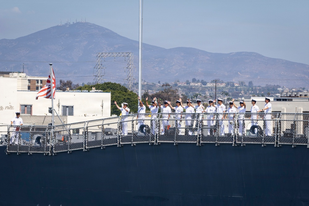 A group of naval personnel dressed in white uniforms are standing along the deck railing of a large warship, with several waving toward the camera or shore. A flag with a red and white design is visible on the left side of the vessel. The background shows a coastal urban area with hills and mountains under a hazy sky.