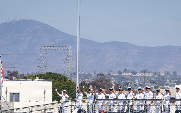 A group of naval personnel dressed in white uniforms are standing along the deck railing of a large warship, with several waving toward the camera or shore. A flag with a red and white design is visible on the left side of the vessel. The background shows a coastal urban area with hills and mountains under a hazy sky.
