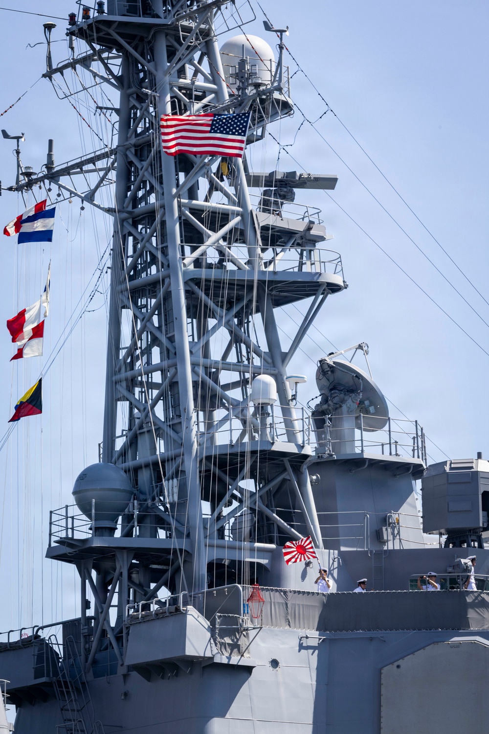 A gray naval warship is shown with its superstructure and mast prominently displayed. Multiple flags are visible including a United States flag and what appears to be the Japanese Maritime Self-Defense Force (JMSDF) Rising Sun naval ensign, suggesting a joint or bilateral naval operation or port visit. Several uniformed personnel are visible on the deck.
