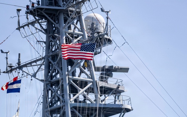 A gray naval warship is shown with its superstructure and mast prominently displayed. Multiple flags are visible including a United States flag and what appears to be the Japanese Maritime Self-Defense Force (JMSDF) Rising Sun naval ensign, suggesting a joint or bilateral naval operation or port visit. Several uniformed personnel are visible on the deck.