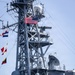 A gray naval warship is shown with its superstructure and mast prominently displayed. Multiple flags are visible including a United States flag and what appears to be the Japanese Maritime Self-Defense Force (JMSDF) Rising Sun naval ensign, suggesting a joint or bilateral naval operation or port visit. Several uniformed personnel are visible on the deck.