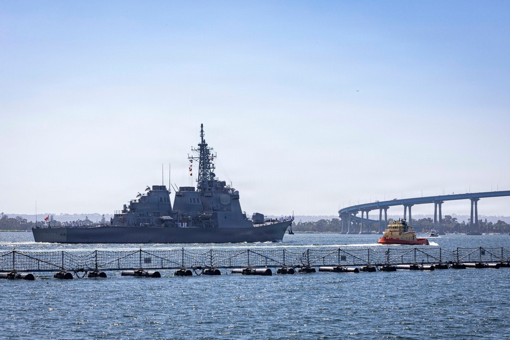 A large gray naval warship is transiting a harbor waterway, accompanied by a bright orange tugboat to its starboard side. In the background, a distinctive curved bridge spans the water, consistent with the Coronado Bridge in San Diego Bay. A floating security barrier or anti-swimmer net is visible in the foreground of the image.