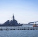 A large gray naval warship is transiting a harbor waterway, accompanied by a bright orange tugboat to its starboard side. In the background, a distinctive curved bridge spans the water, consistent with the Coronado Bridge in San Diego Bay. A floating security barrier or anti-swimmer net is visible in the foreground of the image.