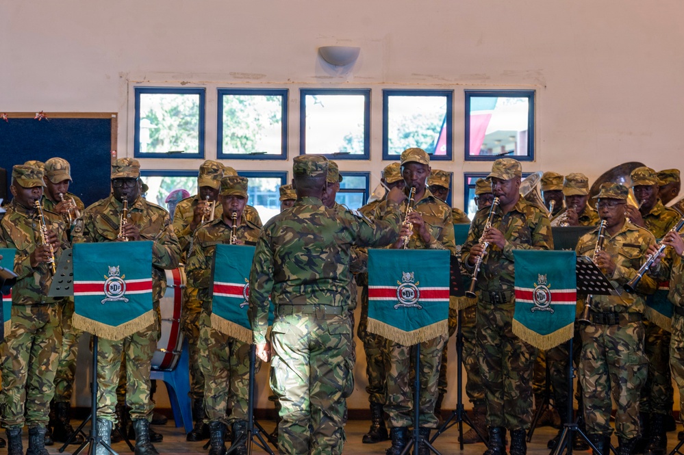 A military band is performing indoors, with musicians in camouflage uniforms playing wind instruments such as clarinets. A conductor stands with his back to the camera, facing and directing the ensemble. Music stands display teal and red pennant banners bearing an emblem, consistent with an East African military organization.