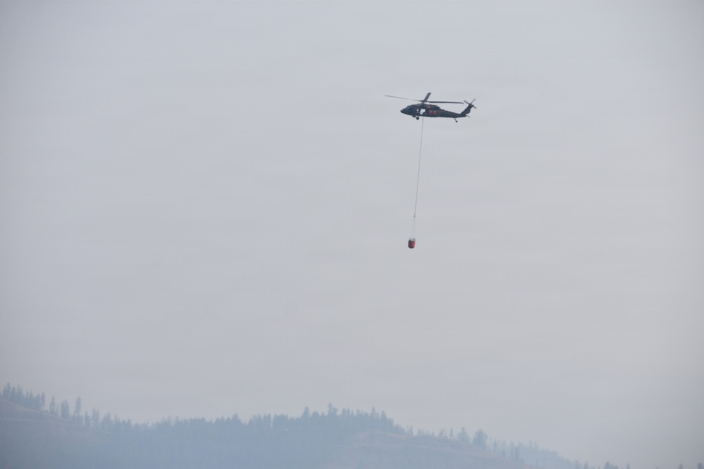A helicopter is flying over a forested, hilly landscape while carrying a suspended water/fire bucket (helibucket) on a long line beneath it. The sky and atmosphere are heavily obscured by smoke or haze, suggesting an active wildfire situation in the area. The scene indicates aerial firefighting operations in progress.