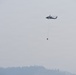 A helicopter is flying over a forested, hilly landscape while carrying a suspended water/fire bucket (helibucket) on a long line beneath it. The sky and atmosphere are heavily obscured by smoke or haze, suggesting an active wildfire situation in the area. The scene indicates aerial firefighting operations in progress.