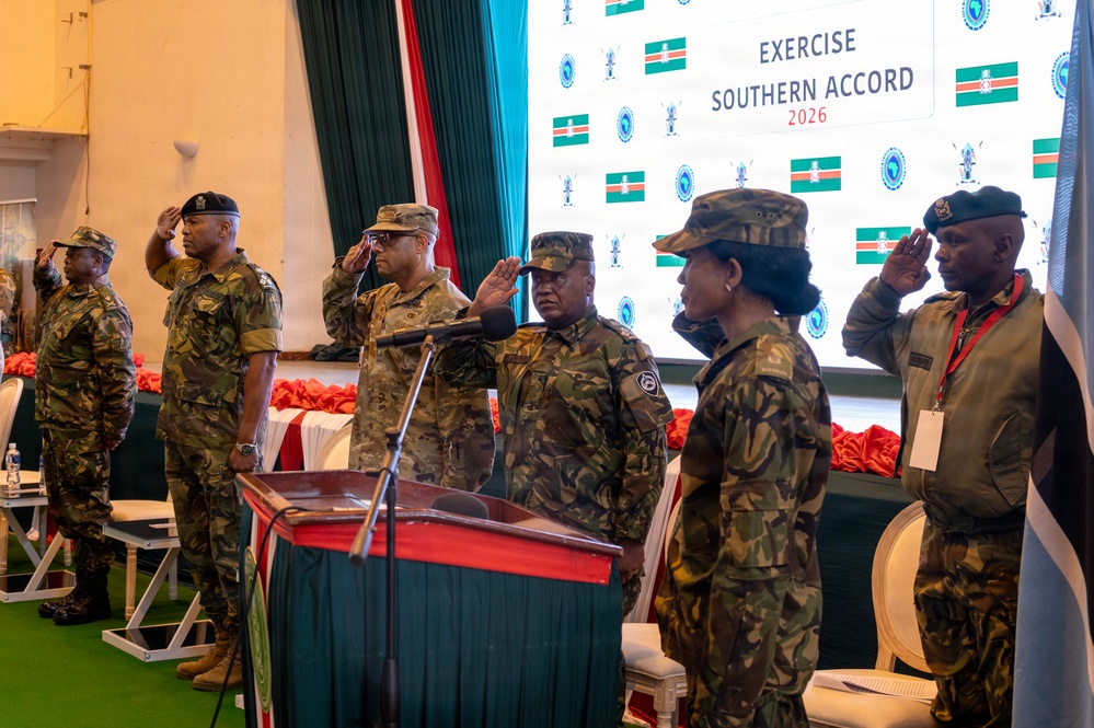 Military personnel from multiple nations are standing at attention and saluting during a formal ceremony for 'Exercise Southern Accord 2026.' The event appears to be an opening or closing ceremony held indoors, with a large branded backdrop displaying flags and the exercise logo. Personnel are wearing a mix of camouflage uniforms and berets, suggesting a multinational military exercise.