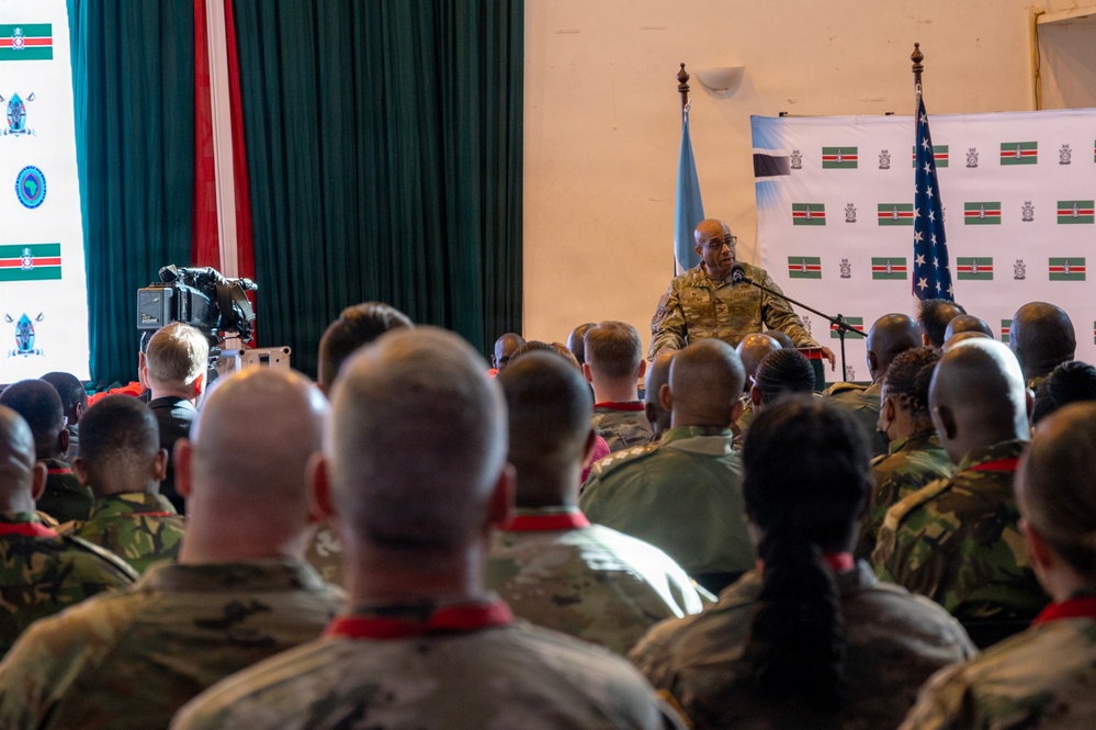 A military briefing or ceremony is taking place in a hall where a senior officer in camouflage uniform addresses a large audience of mixed military personnel. The setting appears to be a joint multinational military event, evidenced by the presence of both a US flag and what appears to be a Suriname flag, along with a UN flag. Personnel are wearing different camouflage uniforms with red lanyards or neckwear.