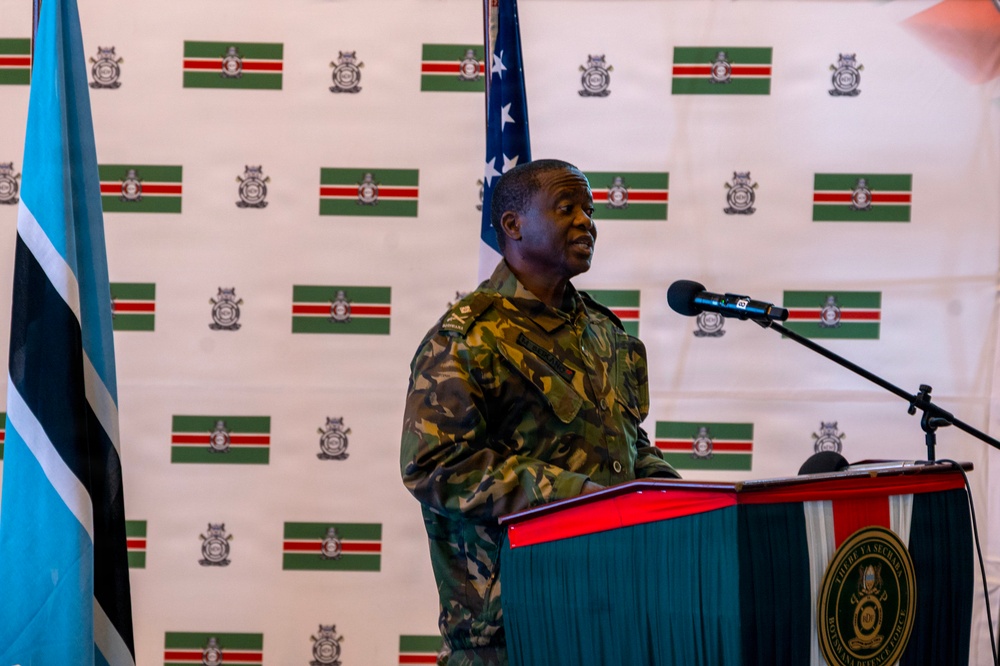 A military officer in camouflage uniform is delivering a speech at a podium with a microphone. The backdrop features a repeated pattern of what appears to be a national flag (green, white, and red stripes with an emblem) alongside the Botswana flag visible on the left. The setting appears to be an official military or government ceremony.