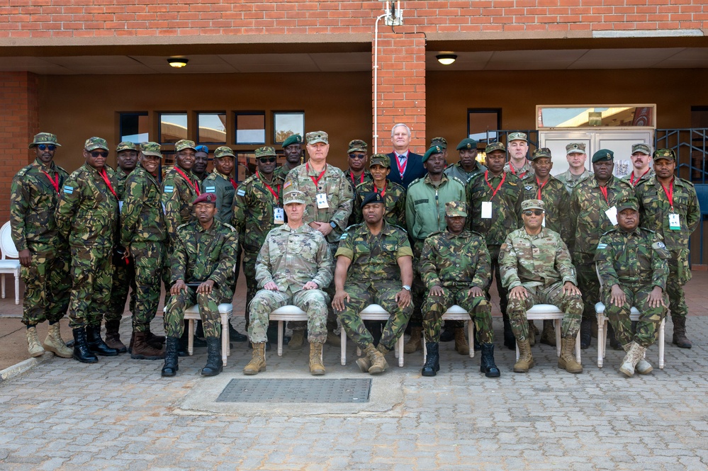 A formal group photograph of approximately 30 military personnel from multiple nations posing in front of a brick building. The group includes soldiers in various camouflage uniforms and one civilian in a suit, suggesting a multinational military exercise, conference, or partnership event. Several participants are wearing red lanyards with identification badges.